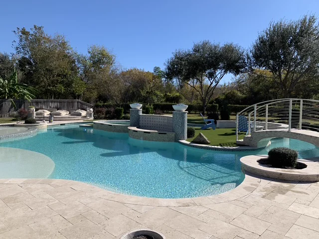 Freeform pool with an arched footbridge, mosaic-tiled raised spa, and travertine deck