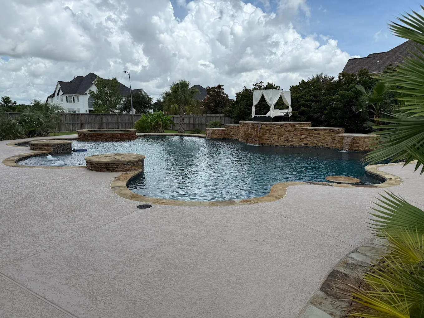 Large freeform pool with a stacked stone waterfall wall, raised spa, and cabana under summer clouds