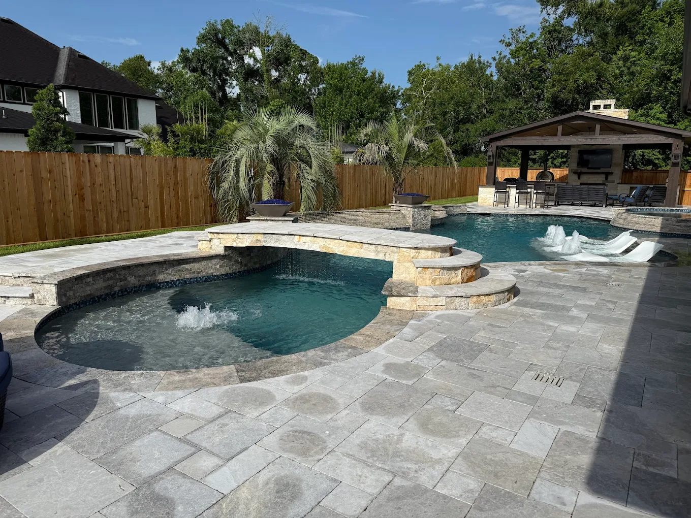 Freeform pool and spa with a stone footbridge, fire bowls, and a covered outdoor kitchen on a travertine deck