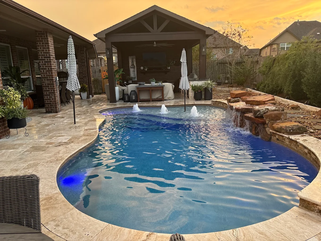 Backyard pool at sunset with a blue underwater light, bubblers, a boulder waterfall, and a covered patio